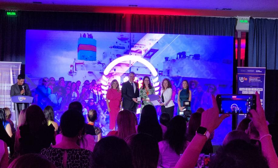 #ParaQuemNaoPodeVer A imagem mostra um evento. Há várias pessoas no palco, algumas segurando microfones, e outras na plateia, algumas tirando fotos com celulares. O ambiente está iluminado com luzes roxas e azuis. No fundo, há um painel com logotipos e textos que não são legíveis nesta imagem.