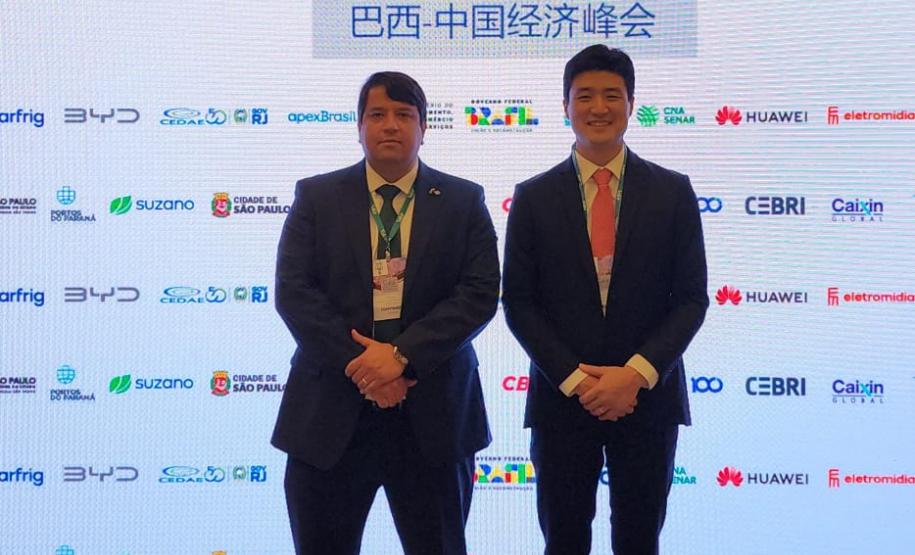 #ParaQuemNaoPodeVer A imagem mostra dois homens de terno em pé, lado a lado, em um evento. O fundo tem um banner com logotipos de várias empresas e as palavras "SUMMIT Valor Econômico BRAZIL-CHINA". Há também caracteres chineses no banner. Os homens estão de frente para a câmera, um deles segurando um microfone.