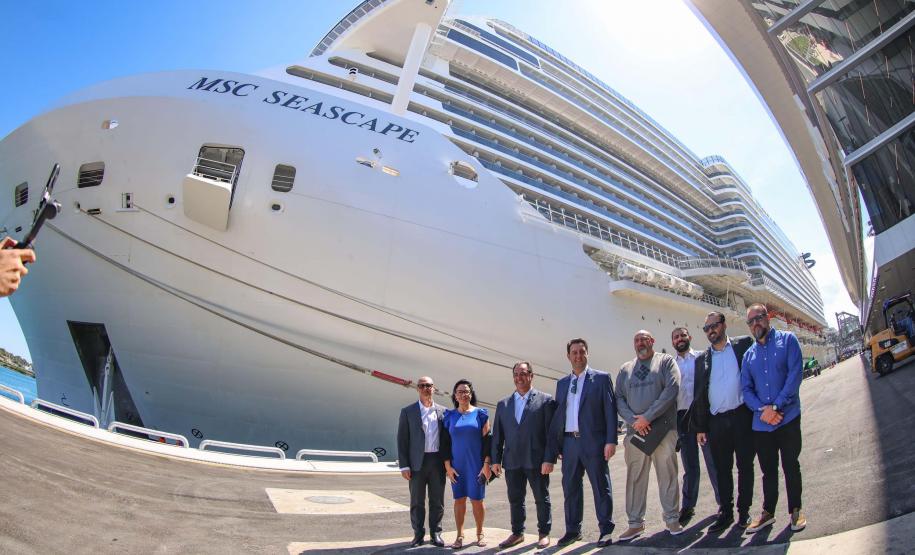 #ParaQuemNaoPodeVer Um grupo de oito pessoas está posando para uma foto em frente a um grande navio de cruzeiro. O navio tem o nome "MSC SEASCAPE" escrito na lateral. As pessoas estão vestidas formalmente, com ternos e camisas sociais. O ambiente parece ser um porto, com o navio atracado ao fundo.