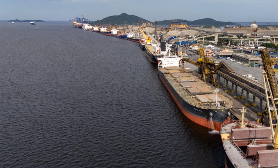 #ParaQuemNaoPodeVer A imagem exibe um porto com um grande navio atracado. O navio está carregado com contêineres e há guindastes ao redor. A água do mar é visível à esquerda, e ao fundo, há uma cidade com colinas.