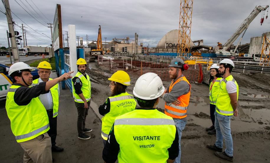 #praquemnaopodever homens e mulheres de colete verde limão estão em uma obra olhando para um rapaz de capacete branco que aponta ao longe. O céu está cinza, o chão de barro está molhado. Ao fundo, máquinas estão suspensas por causa da chuva que dá sinais de que irá voltar