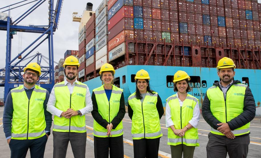 #ParaQuemNaoPodeVer Seis pessoas estão posando para uma foto em frente a um cenário de contêineres empilhados em um porto. Todos estão usando coletes de segurança amarelos e capacetes amarelos. Ao fundo, há uma estrutura de guindaste azul e os contêineres têm várias cores e logotipos, incluindo "MAERSK".
