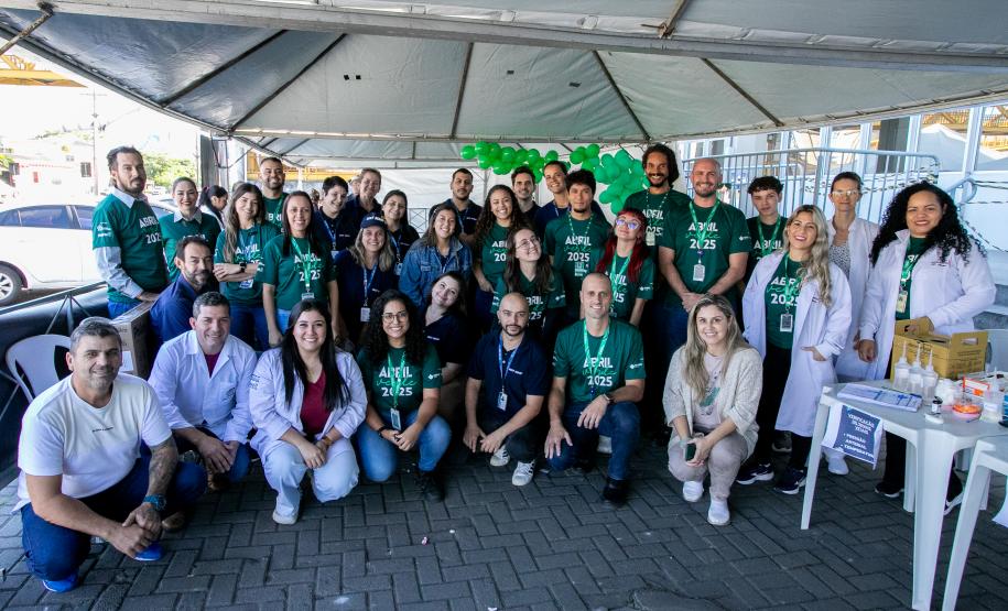#ParaQuemNaoPodever Um grupo grande de pessoas está posando para uma foto em um ambiente externo. A maioria delas está usando camisetas verdes com a inscrição "ABRIL VERDE 2025". Algumas pessoas estão agachadas na frente, enquanto outras estão em pé atrás. Há uma tenda ao lado direito da imagem e algumas bexigas verdes. No canto inferior esquerdo, há uma mesa branca com alguns objetos em cima, incluindo um livro amarelo e um panfleto azul.