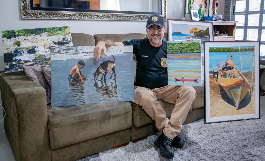 #ParaQuemNaoPodeVer Homem sentado em um sofá segurando duas pinturas. Ele está usando uma camiseta preta e um boné azul com um emblema. As pinturas mostram cenas de praia: uma com duas pessoas e um cachorro na água, e outra com um barco na areia. Ao lado dele, há mais pinturas e uma moldura decorativa na parede. O ambiente parece ser uma sala de estar, com um sofá bege e uma mesa com objetos ao fundo.