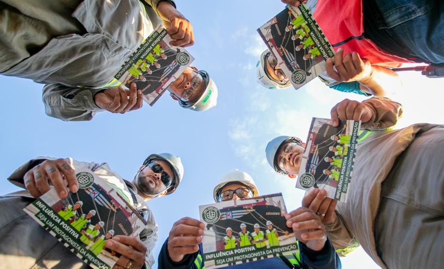 #ParaQuemNaoPodeVer Imagem exibe várias pessoas em um círculo, olhando para a câmera de baixo para cima. Elas estão segurando panfletos ou revistas que têm a imagem de trabalhadores de construção civil na capa. O texto na capa diz: "INFLUÊNCIA POSITIVA: ESSA É A POSTURA DA PREVENÇÃO". As pessoas na foto estão usando capacetes de segurança e óculos de proteção, sugerindo que são trabalhadores de uma obra ou indústria.