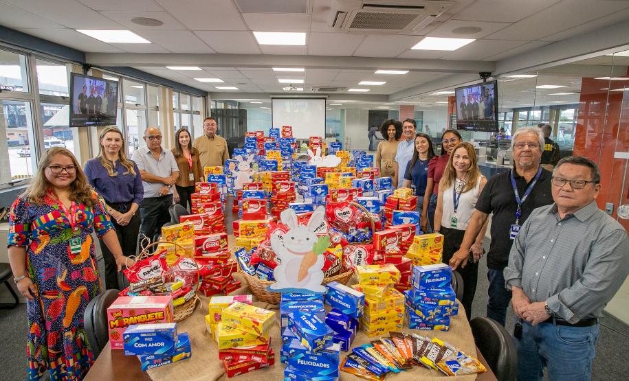 #ParaQuemNaoPodeVer Imagem exibe um grupo de pessoas em um escritório. Na imagem, há uma mesa cheia de pacotes de biscoitos e outros alimentos embalados. As pessoas estão sorrindo e posando para a foto, e parecem estar participando de algum tipo de evento ou campanha de arrecadação de alimentos.
