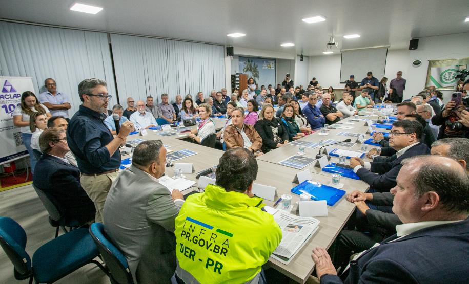 #praquemnaopodever setenta pessoas estão sentadas olhando para um senhor em pé, de camisa azul escura e calça bege. Ele segura um microfone na mão e com olhar focado por trás dos óculos, observa o público que o acompanha. A sala está com as cortinas brancas fechadas e ao fundo a imprensa grava o encontro.
