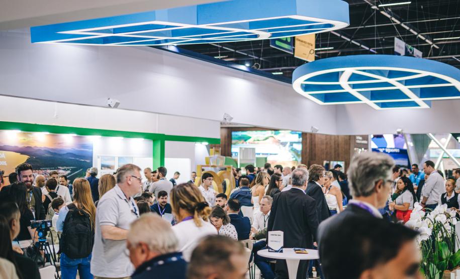 #ParaQuemNaoPodeVer Foto exibe um evento ou feira com muitas pessoas. Há uma estrutura grande e moderna no teto, com formas geométricas azuis. As pessoas estão em pé e algumas estão sentadas em mesas, parecendo interagir ou observar algo. O ambiente está movimentado e há uma variedade de pessoas, algumas com mochilas.