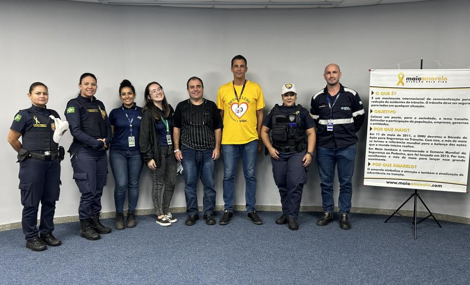 #ParaQuemNaoPodeVer Foto de um grupo de oito pessoas, sendo quatro delas usando uniformes de agentes de trânsito ou policiais, e as outras quatro usando roupas casuais. Eles estão em pé, lado a lado, em frente a uma parede clara e um tapete azul. À frente do grupo, há um banner com informações sobre o movimento Maio Amarelo, que é uma campanha de conscientização para a segurança no trânsito. O banner tem detalhes em amarelo e branco, com o símbolo do laço amarelo. Todos estão sorrindo e olhando para a câmer