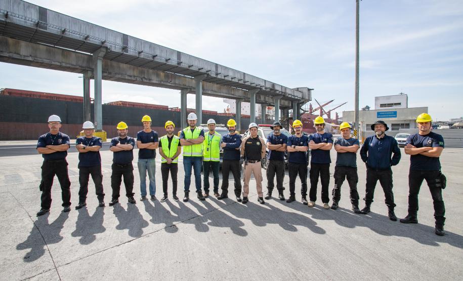 #ParaQuemNaoPodeVer Foto de um grupo de pessoas em um porto. Elas estão alinhadas, usando capacetes de segurança amarelos e brancos, e algumas vestem coletes refletivos. Ao fundo, há uma estrutura de concreto que de carregamento de granéis.