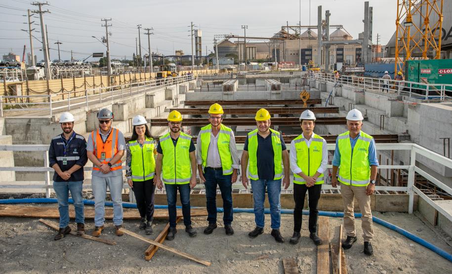 #ParaQuemNaoPodeVer Grupo de oito pessoas em um canteiro de obras. Todos estão usando equipamentos de segurança, como capacetes e coletes refletivos. A maioria veste coletes amarelos, enquanto uma pessoa usa um colete laranja. O cenário ao fundo mostra estruturas de concreto e uma grua amarela, indicando que estão em uma área de construção. O céu está nublado e há postes de eletricidade ao fundo.
