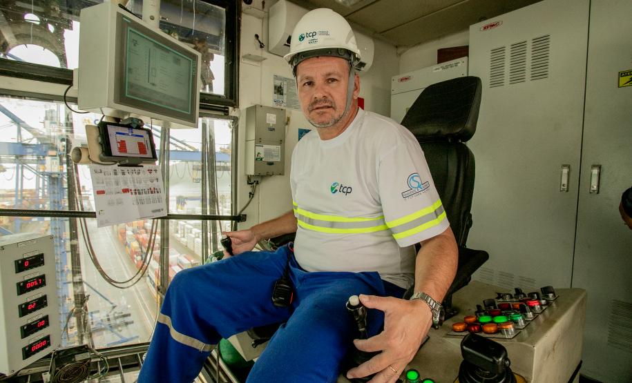 #ParaQuemNaoPodeVer Foto de um homem sentado em uma cabine de operação. Ele está usando capacete branco com o logotipo da empresa "ICTSI" e uniforme azul com faixas refletivas amarelas. O homem está sentado em uma cadeira diante de um painel de controle com vários botões coloridos e alavancas. À frente dele, há uma tela de computador com gráficos e informações técnicas. A cabine tem janelas grandes, permitindo ver estruturas metálicas e o que parece ser um terminal portuário ao fundo. O ambiente sugere que