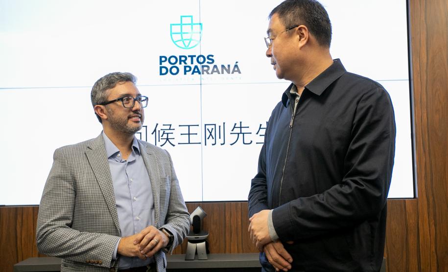 #praquemnaopodever na foto, dois homens conversam em frente a uma tela branca com logo escrita "Portos do Paraná" e dizeres em mandarim embaixo. A esquerda, o homem branco de terno cinza, camisa azul e óculos segura as mãos enquanto conversa com um homem chinês mais alto, de óculos, camisa e calça pretas.