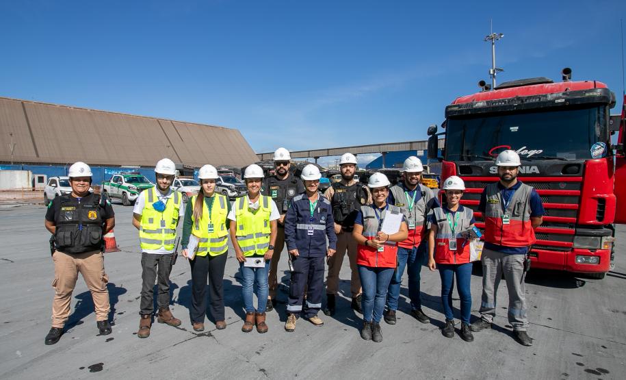 #ParaQuemNaoPodeVer Imagem mostra grupo de pessoas posando em frente a um caminhão vermelho. Todos estão usando capacetes de segurança e coletes refletivos, indicando que podem estar em um ambiente de trabalho ou construção. Ao fundo, há um grande galpão e o céu está azul.