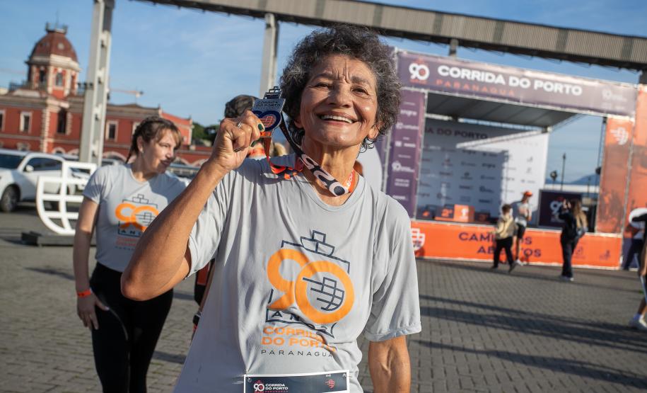 #praquemnaopodever na foto, ao centro, idosa com camisa cinza e letras em laranja escrito 90 Portos do Paraná, ergue com um sorriso no rosto a medalha recebida após concluir a Corrida do Porto.