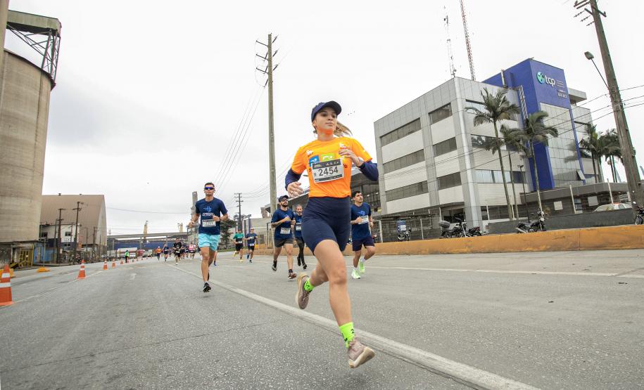 #praquemnaopodever na foto aparece no centro uma mulher correndo com olhar para distraído para frente. Calça um tennis cinza com meias verde-limão, camisa laranja com o número 2454e usando bermuda colada azul e viseira azul. Ao fundo a direita está uma empresa azul e cinza e atras dela aparecem outros atletas correndo
