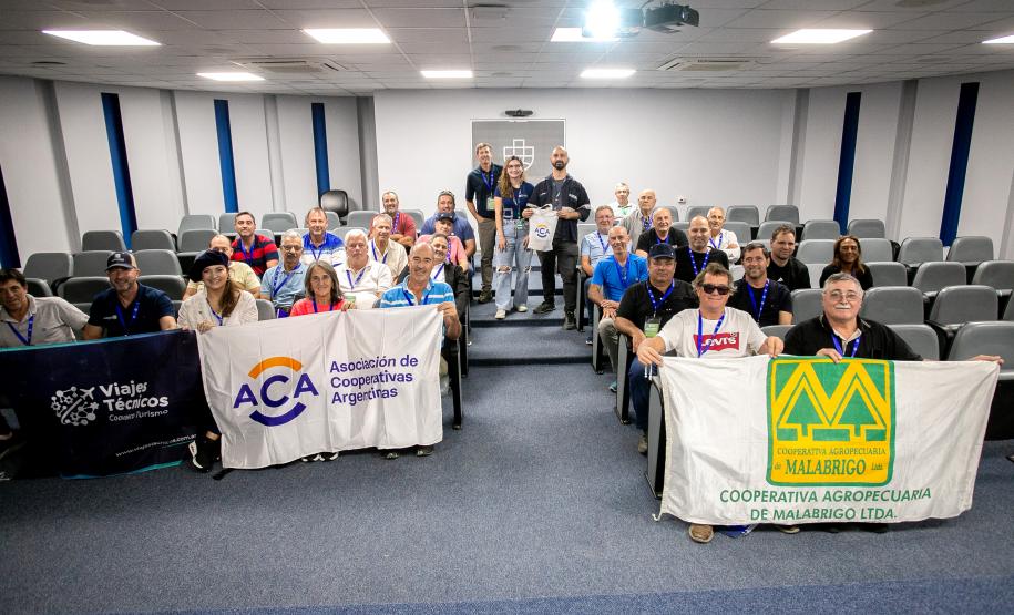 #ParaQuemNaoPodeVer Foto de um grupo de pessoas sentadas em cadeiras de auditório, posando para a câmera. Elas estão sorrindo e segurando três faixas diferentes. A faixa do topo, à esquerda, tem o texto “Viajes Teóricos” e “Colectivo”, a faixa do meio tem o logotipo da ACA e diz “Asociación de Cooperativas Argentinas”, e a faixa da frente, em destaque, tem um grande “M” amarelo e verde e o texto “Cooperativa Agropecuária de Malabrigo Ltda.”. O ambiente parece ser uma sala de reuniões ou auditório, com cadei