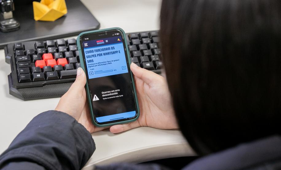 #ParaQuemNaoPodeVer Imagem mostra uma foto de uma pessoa segurando um celular. O celular está exibindo uma página da internet com o título “COMO FUNCIONAM OS GOLPES POR WHATSAPP E SMS”. Abaixo do título, há um texto menor, mas não está totalmente legível na imagem. O celular está em cima de um teclado de computador com algumas teclas vermelhas e outras pretas. No canto superior direito da mesa, há um objeto amarelo que parece ser uma dobradura de papel (origami). A pessoa está usando uma blus
