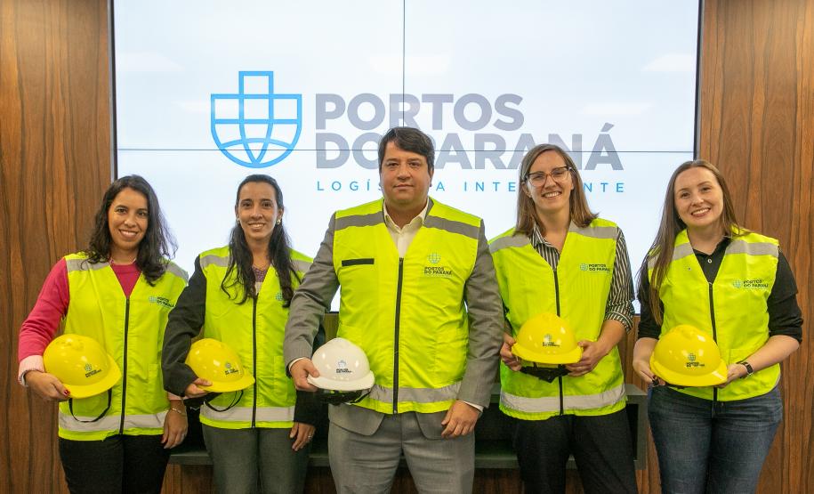 #ParaQuemNaoPodeVer Foto de cinco pessoas em pé, todas usando coletes de segurança amarelo e segurando capacetes (a maioria amarelos, um branco). Elas estão sorrindo e posando para a foto em frente a um painel com o logotipo azul e as palavras “PORTOS DO PARANÁ LOGÍSTICA INTELIGENTE”. O grupo é composto por quatro mulheres e um homem, todos com aparência profissional.