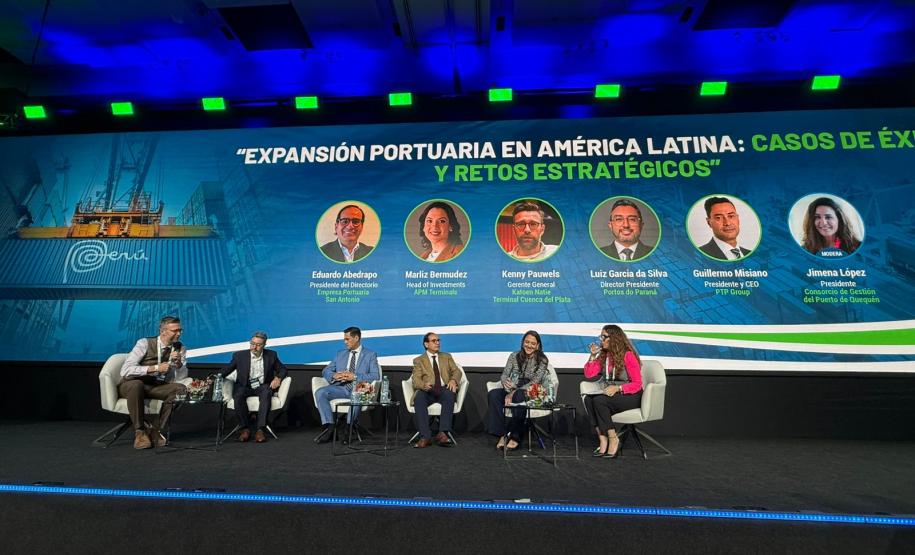 #ParaQuemNaoPodeVer Palco de conferência com seis pessoas sentadas em cadeiras, participando de um painel de discussão. Ao fundo, há um grande painel azul com a imagem de um contêiner portuário e o título do evento em espanhol: "Expansión portuaria en América Latina: casos de éxito y retos estratégicos". Abaixo do título, aparecem fotos e nomes dos participantes do painel, com seus respectivos cargos e empresas. O ambiente é formal, com todos vestidos de maneira profissional. O palco está bem iluminado e há