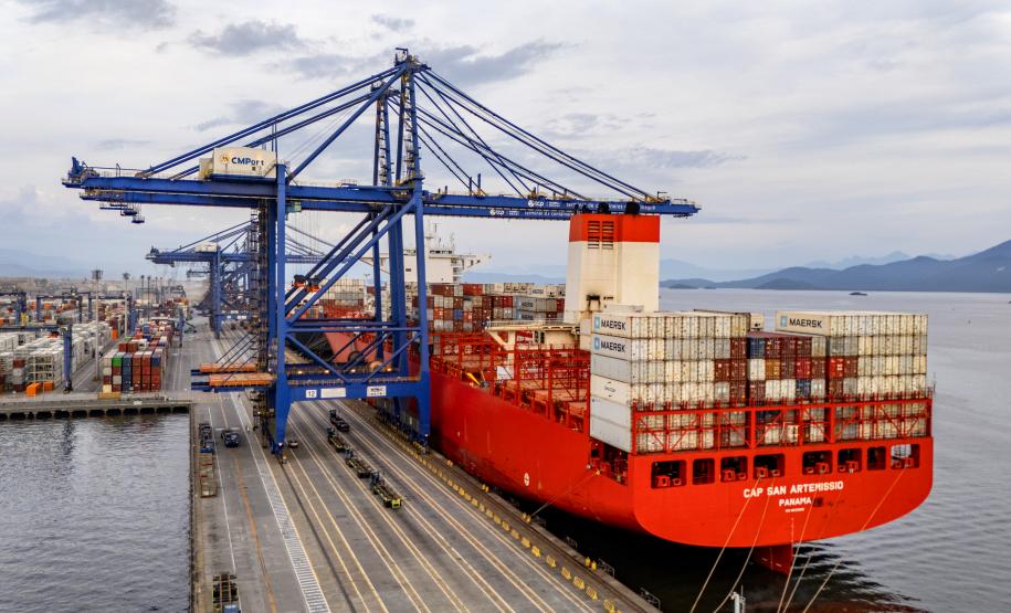 #ParaQuemNaoPodeVer Imagem de um navio cargueiro vermelho atracado em um porto. O navio está carregado com vários contêineres empilhados e há guindastes azuis grandes posicionados para carregar ou descarregar os contêineres. À esquerda, é possível ver parte do cais com caminhões e veículos pequenos. O mar está calmo e o céu está parcialmente nublado.