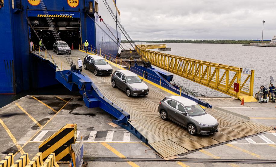 #ParaQuemNaoPodeVer A imagem mostra uma rampa amarela de acesso à balsa, que está ancorada em um porto. Há cinco carros visíveis, alinhados e entrando na balsa, que é azul. No fundo, vê-se o rio e parte da margem oposta. Sinais circulares amarelos na entrada da balsa indicam altura máxima de 7,5 metros e largura máxima de 5,2 metros. O ambiente parece ser de um terminal portuário.