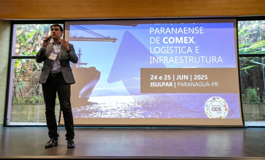#ParaQuemNaoPodeVer Foto de um homem em pé, falando ao microfone em um palco. Ele está usando calça preta, sapato preto, camisa clara e blazer escuro. Atrás dele há um telão grande com a seguinte mensagem:  "PARANAENSE DE COMEX LOGÍSTICA E INFRAESTRUTURA 24 e 25 JUN 2025 ISUPAR, PARANAGUÁ-PR"  No canto inferior esquerdo do telão há um selo colorido com os dizeres "ODS 2030". Ao fundo, através de uma janela de vidro, é possível ver plantas e uma parede de pedras.