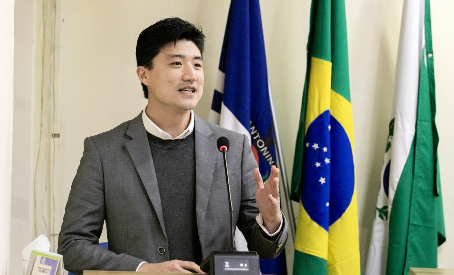 #ParaQuemNaoPodeVer foto de um homem jovem, de pele clara e cabelo preto, vestindo terno cinza, suéter escuro e camisa branca. Ele está em pé atrás de um púlpito, falando ao microfone, com expressão sorridente. Atrás dele, há bandeiras, incluindo a do Brasil e outra azul com a palavra "NITOINI" parcialmente visível. O ambiente parece ser formal, possivelmente uma apresentação ou discurso.