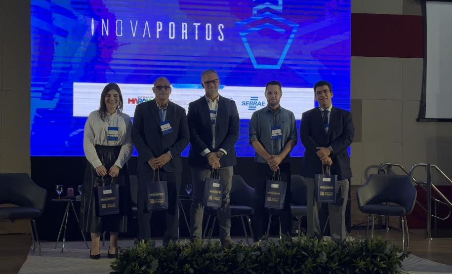 #ParaQuemNaoPodeVer Na imagem do evento, há cinco pessoas (quatro homens e uma mulher) em pé, posando para foto em um palco. Todos estão vestidos de forma formal, com crachás pendurados no pescoço. Eles seguram sacolas pretas com o logo do evento. Ao fundo, há um painel azul grande com a palavra "INOVAPORTOS" escrita na vertical. Também é possível ver algumas cadeiras vazias no palco e plantas decorativas na frente. O ambiente parece ser de uma conferência ou seminário.