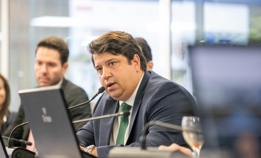 #praquemnaopodever foto mostra homem de terno azul, camisa branca e gravata verde sentado olhando para além da câmera e falando ao microfone. Ao fundo, estão desfocados os colegas que prestam atenção ao homem moreno e olhar centrado.