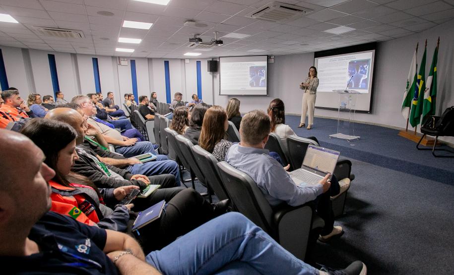 #ParaQuemNaoPodeVer Auditório com várias pessoas sentadas em cadeiras, assistindo a uma apresentação. No fundo da sala, uma mulher está em pé, falando ao microfone, com um telão atrás dela exibindo um slide. Há bandeiras do Brasil e de outros estados ou instituições ao lado do palco. As pessoas na plateia parecem estar prestando atenção, algumas com crachás e blocos de anotações. O ambiente é iluminado por luzes de teto e tem paredes brancas com detalhes em azul.