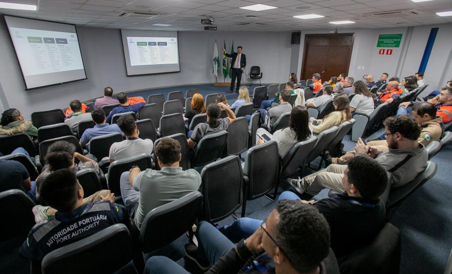 #ParaQuemNaoPodeVer Sala de aula ou auditório com várias pessoas sentadas em cadeiras, todas voltadas para a frente, assistindo a uma apresentação. Na frente da sala, há uma pessoa de pé, aparentemente palestrando, e dois telões com slides projetados na parede. No canto direito, há três bandeiras, incluindo a do Brasil. Muitas pessoas estão usando camisetas cinza com a inscrição “AUTORIDADE PORTUÁRIA PORT AUTHORITY” nas costas. O ambiente parece formal e voltado para treinamento ou reunião.