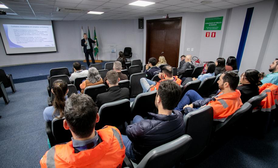 #ParaQuemNaoPodeVer Sala de reuniões vista de trás, com várias pessoas sentadas em cadeiras, assistindo a uma apresentação. Muitas dessas pessoas estão usando uniformes laranja com faixas refletivas, típicos de trabalhadores de áreas industriais ou de segurança. Na frente da sala, há uma pessoa em pé, aparentemente apresentando um slide projetado na parede. Ao lado do apresentador, estão duas bandeiras (provavelmente do Brasil e do estado). A sala tem piso azul, paredes claras e teto com iluminação embutida