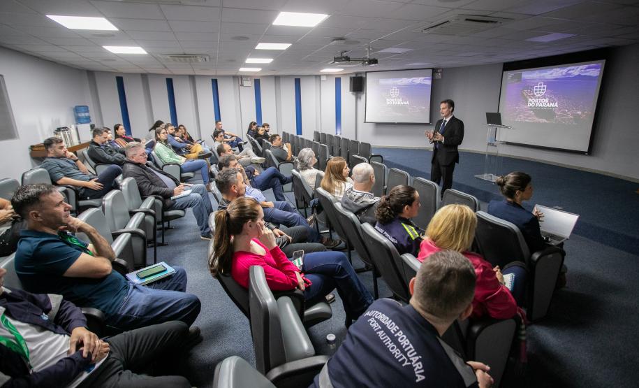 #ParaQuemNaoPodeVer Auditório com várias pessoas sentadas em cadeiras, todas de cabeça baixa. Na frente, um homem de terno está em pé, também de cabeça baixa, próximo a um púlpito transparente. Ao fundo, há dois telões projetando a mesma imagem, com fundo roxo e texto branco, mas o texto não está totalmente legível na foto. O ambiente é fechado, com iluminação de teto e paredes claras.