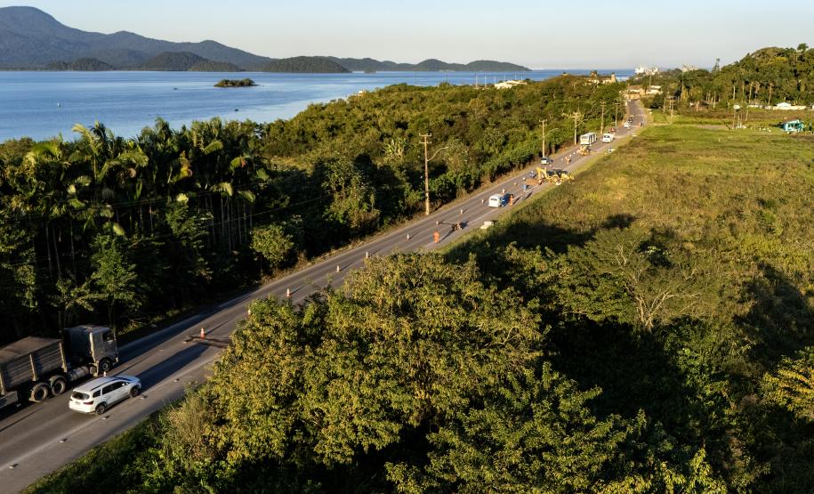 #ParaQuemNaoPodeVer Foto aérea de uma estrada rodeada por vegetação densa e árvores. À esquerda da imagem, há um caminhão e um carro branco na estrada. À direita, é possível ver um corpo d’água azul (provavelmente um rio ou mar) e montanhas ao fundo. A estrada corta a paisagem diagonalmente, e há postes de energia acompanhando o trajeto.