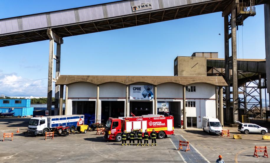 #ParaQuemNaoPodeVer foto de um pátio industrial ou de uma garagem de veículos grandes. No centro da imagem, há dois caminhões: um caminhão de bombeiros vermelho com a inscrição "BOMBEIROS" e outro caminhão branco com detalhes azuis, que parece ser de resgate ou suporte. Ambos estão estacionados próximos a um prédio de estrutura robusta, com cobertura alta e colunas de concreto. Há cones e barreiras laranja ao redor dos veículos, indicando uma área isolada. Algumas pessoas com coletes refletores estão próxi
