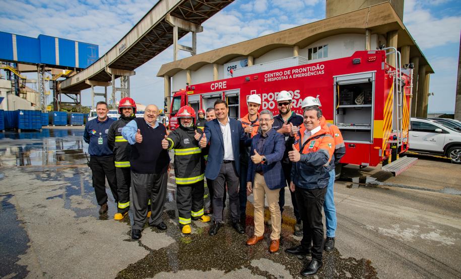 #praquemnaopodever Na foto um grupo de homens faz o sinal de jóia com o polegar e sorriem em frente a um caminhão de bombeiros. Ao fundo, um predio branco e dalas suspensas que conectam empresas ao cais.