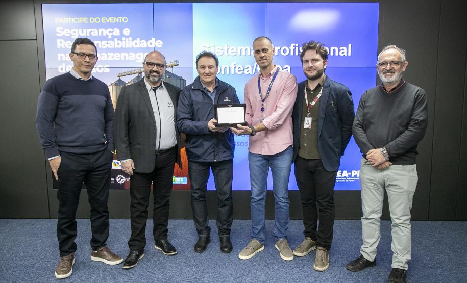 #ParaQuemNaoPodeVer Foto de seis homens posando juntos em frente a um painel azul. Eles estão em pé, lado a lado, sorrindo para a câmera. Um dos homens, no centro, segura um objeto retangular, que parece ser um prêmio ou uma placa. O painel atrás deles tem textos em português, incluindo frases como “PARTICIPE DO EVENTO”, “Segurança e Responsabilidade no Armazenagem”, e “Sistema Profissional”. Todos estão vestidos de maneira casual ou social, com camisas, blazers e calças.