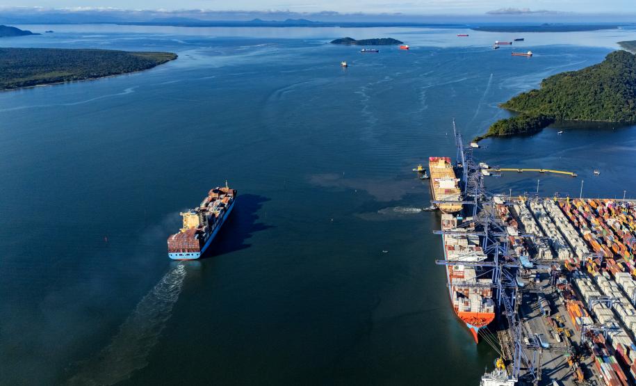 #ParaQuemNaoPodeVer Imagem aérea de um porto mostrando um grande navio saindo do cais, cercado de contêineres e equipamentos portuários. O mar, de azul intenso, ocupa a maior parte da imagem, com algumas pequenas ilhas verdes ao fundo. Outros navios menores podem ser vistos mais afastados, navegando ao longe.