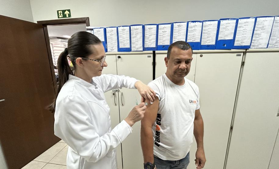 #ParaQuemNaoPodeVer Cena dentro de um consultório ou ambiente de saúde. Uma mulher de cabelo preso, óculos e jaleco branco está aplicando uma injeção no braço de um homem. O homem veste uma camiseta branca com a palavra "ENFERMAGEM" estampada. Eles estão em pé, próximos a armários brancos, com vários papéis presos em pastas azuis na parede ao fundo. Há também uma placa de saída de emergência verde na parede superior direita.