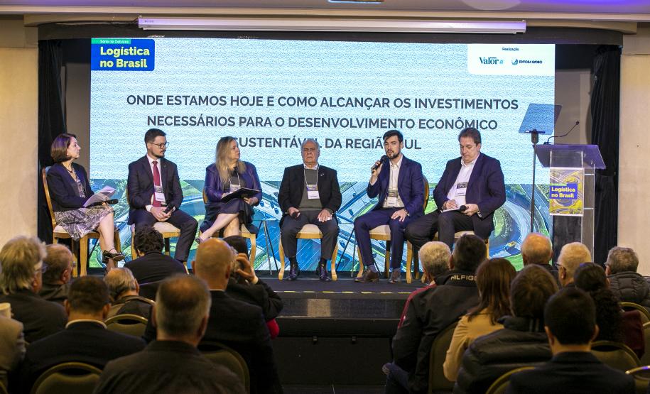 #ParaQuemNaoPodeVer Foto de um painel em um evento corporativo, com cinco pessoas (quatro homens e uma mulher) sentadas no palco, participando de um debate. No fundo, há um grande telão com o texto: "Logística no Brasil ONDE ESTAMOS HOJE E COMO ALCANÇAR OS INVESTIMENTOS NECESSÁRIOS PARA O DESENVOLVIMENTO ECONÔMICO SUSTENTÁVEL DA REGIÃO SUL". O público está sentado de frente para o palco, acompanhando a discussão. No canto inferior direito da tela, há um púlpito transparente com a mesma identidade visual do