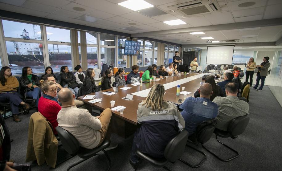 #ParaQuemNaoPodeVer Foto mostra uma reunião em uma sala de conferências. Ao redor de uma mesa grande e retangular, há muitas pessoas sentadas, a maioria prestando atenção em alguém que está falando ou apresentando (próximo a um quadro de projeção na ponta da mesa). A sala tem janelas grandes ao fundo, por onde é possível ver navios. As pessoas na reunião são de perfis variados e algumas estão com laptops ou papéis na mesa. A imagem sugere um ambiente corporativo ou de trabalho colaborativo.