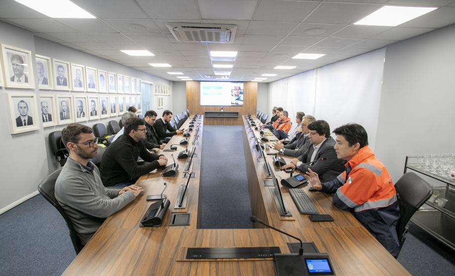 #ParaQuemNaoPodeVer Sala de reuniões com uma mesa longa de madeira, onde diversas pessoas estão sentadas dos dois lados, aparentemente participando de uma reunião. Algumas pessoas usam uniformes laranja e cinza, que parecem ser de alguma empresa ou serviço. Há microfones e pequenos dispositivos em frente a cada pessoa na mesa, sugerindo que é uma reunião formal ou corporativa. Na parede à direita estão pendurados vários quadros com fotos de pessoas, provavelmente fotos de ex-diretores, presidentes ou homena