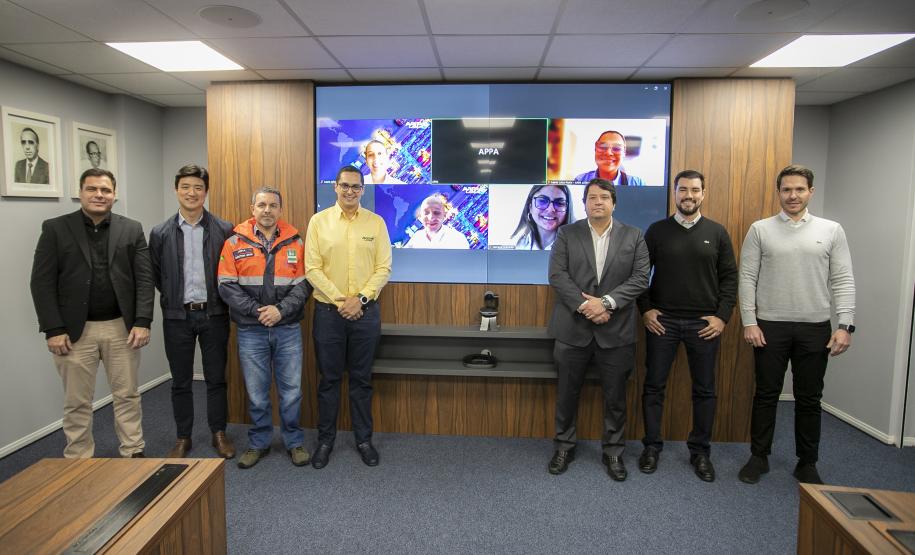 #ParaQuemNaoPodeVer Foto de sete homens em uma sala de reunião corporativa. Eles estão todos em pé, lado a lado, olhando para a câmera. Quatro deles estão vestidos de forma casual e três usam roupas mais formais (terno ou camisa social). Dois dos homens ao centro estão utilizando uniformes: um de jaqueta laranja e outro de jaqueta amarela, que parecem ser de alguma empresa ou serviço. Atrás deles, há uma televisão grande na parede exibindo uma imagem colorida com pessoas em uma chamada de vídeo, sugerindo q