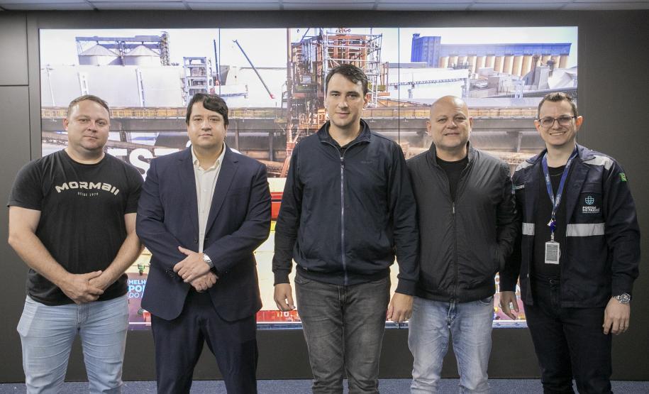 #ParaQuemNaoPode Ver Imagem mostra cinco homens posando para uma foto em grupo. Eles estão em pé, lado a lado, com um fundo que parece ser uma imagem de uma área industrial ou de construção, com estruturas metálicas e guindastes. Um dos homens, na parte superior esquerda, veste uma camiseta preta com a palavra "Mormaii" escrita. Outro homem, ao lado dele, está de terno azul escuro. Os demais usam roupas casuais ou jaquetas, e um deles, na parte inferior, usa um crachá e uniforme preto com detalhes