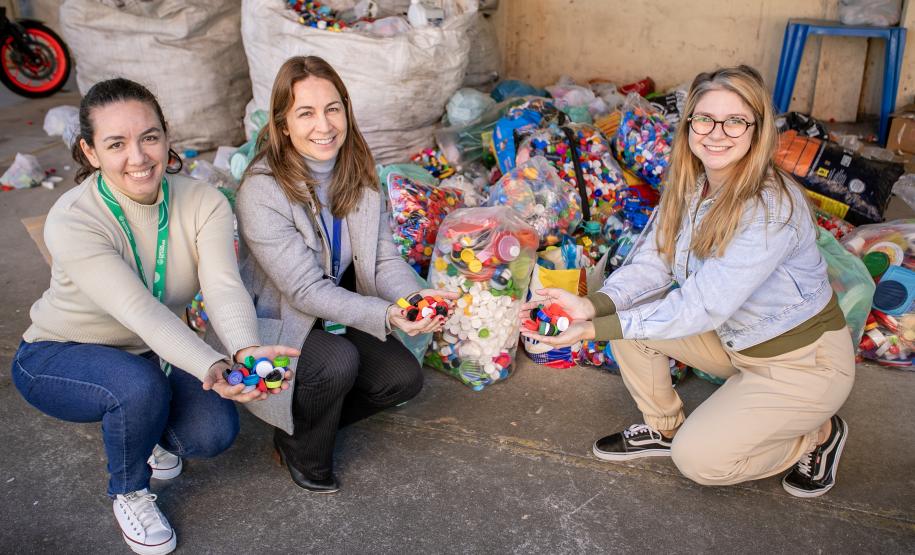 #ParaQuemNaoPodeVer Foto de três mulheres sorrindo, agachadas ou ajoelhadas próximas umas das outras. Elas estão segurando várias tampinhas plásticas coloridas nas mãos. Ao fundo, há vários sacos grandes cheios de tampinhas plásticas, sugerindo uma campanha de reciclagem ou coleta de tampas. O ambiente parece ser um galpão ou área de armazenamento. As três estão vestidas de forma casual e aparentam estar felizes com a atividade.