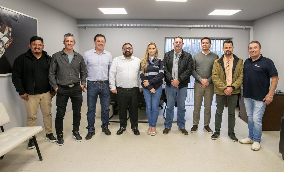 #ParaQuemNaoPodeVer Foto mostra um grupo de nove pessoas, todas em pé e organizadas em fila. O ambiente parece um escritório, com paredes claras, piso claro e janelas atrás do grupo. As pessoas estão vestidas de maneira casual a social, com camisas, jaquetas, calças jeans e algumas de camiseta. Há uma mulher no grupo enquanto o restante do grupo é composto por homens. Todos posam para a foto sorrindo.