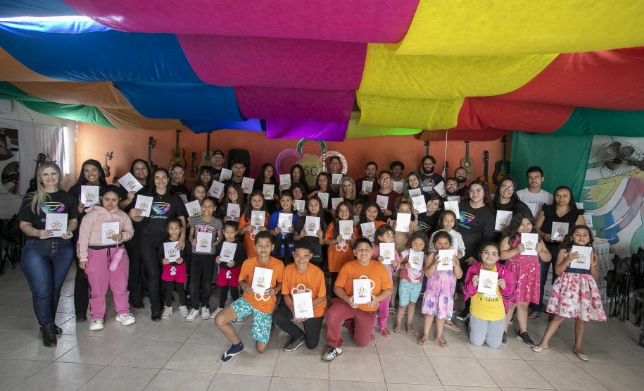 #praquemnaopodever foto mostra um grupo com 50 pessoas segurando um livro com capa branca posando para a foto. Estão em uma sala com parede laranja ao fundo e panos coloridos no teto. Ao fundo, uma um desenho de rosa com o nome 5C ao centro.