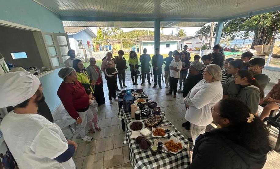 #praquemnaopodever foto mostra um grupo de pessoas em círculo em volta de uma mesa com toalha xadrez e repleta de comida. A maioria olha para uma pessoa que fala no outro lado da foto.
