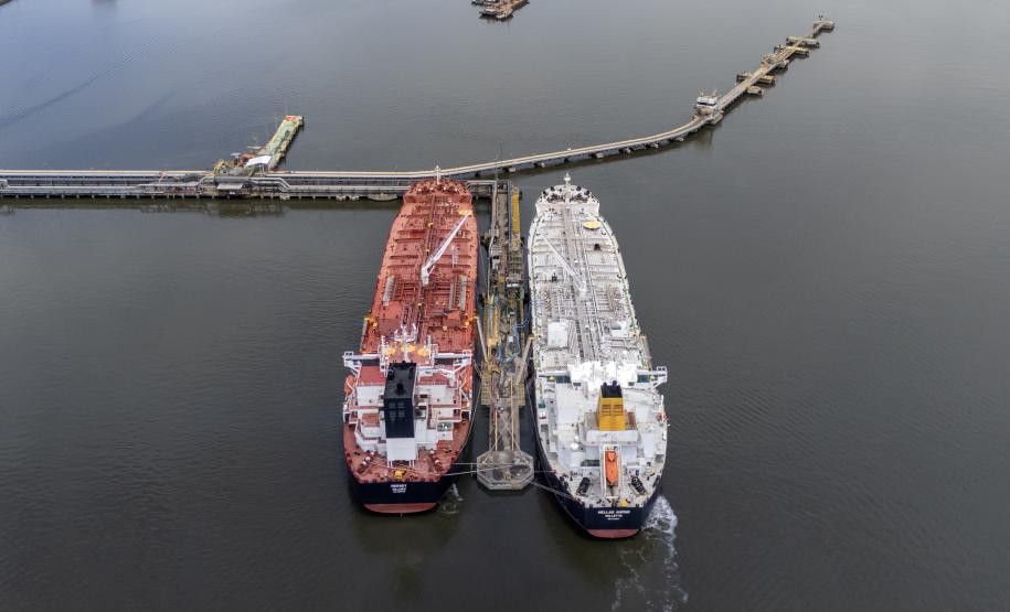 #ParaQuemNaoPodeVer Foto aérea de dois navios-tanque atracados lado a lado em um píer no mar. O navio da esquerda é vermelho e o da direita é branco com detalhes em amarelo. Ambos estão alinhados paralelamente e conectados ao píer, que se estende da margem até o local onde eles estão. Próximo ao píer, à direita, há um pequeno barco navegando.
